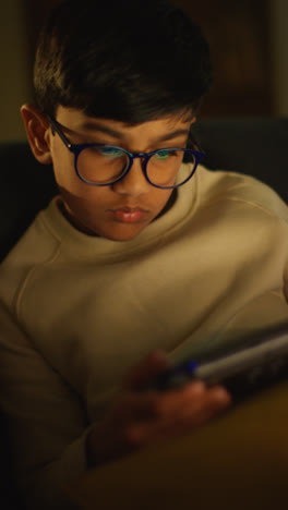 Vertical-Video-Close-Up-Of-Young-Boy-Sitting-On-Sofa-At-Home-Playing-Game-Or-Streaming-Onto-Handheld-Gaming-Device-At-Night